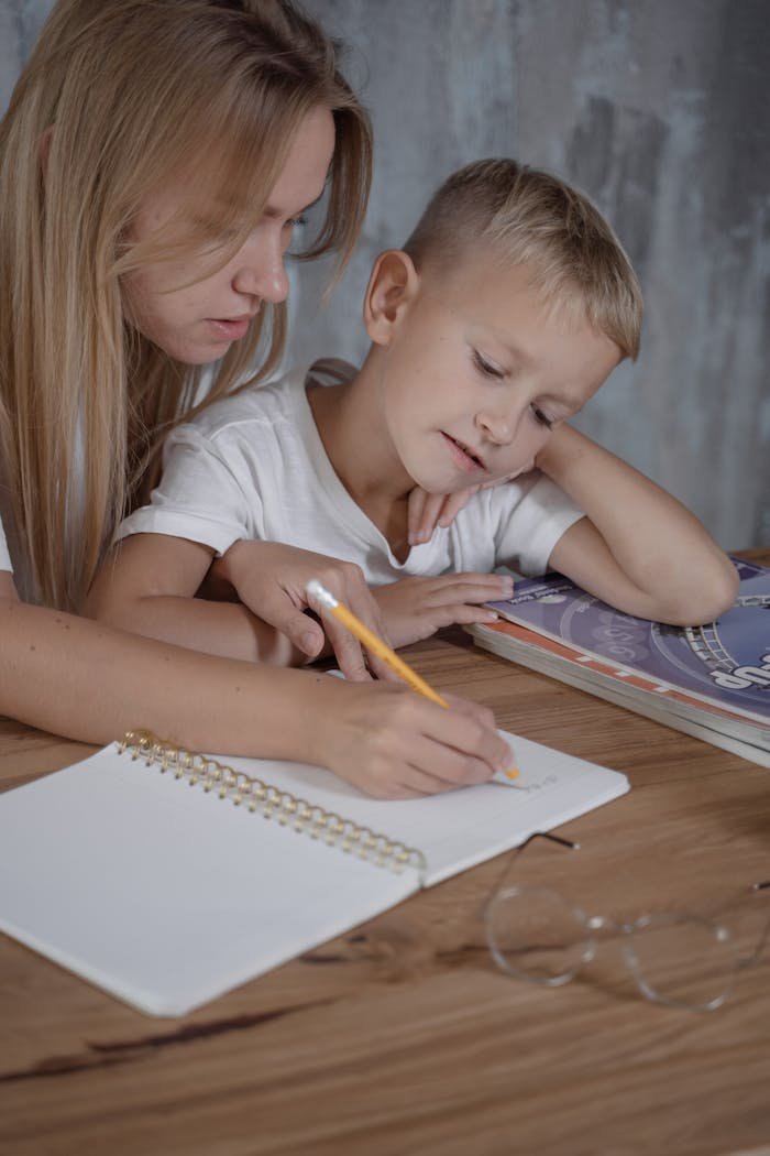 A mother assists her son with his homework, writing in a notebook placed on a wooden surface.