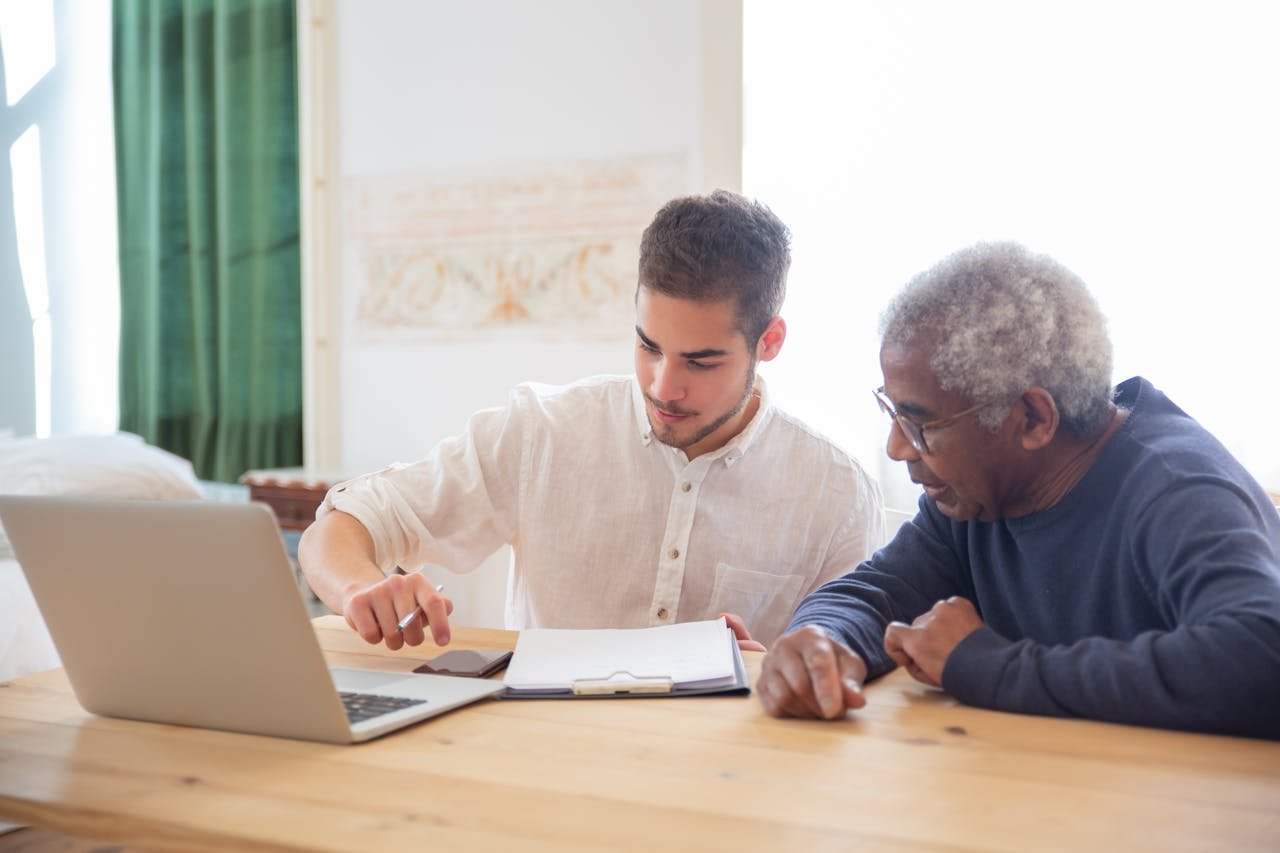 Young man teaches elderly gentleman to use technology at home.